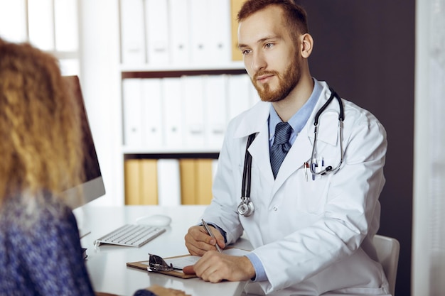 Sympathique médecin à barbe rouge et patiente discutant de l'examen de santé actuel tout en étant assis dans une clinique ensoleillée. Notion de médecine.