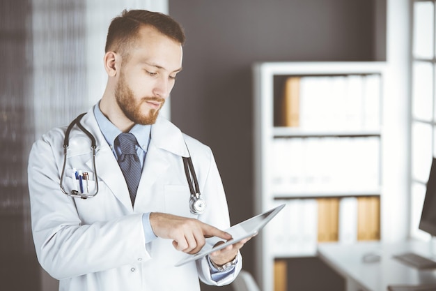 Sympathique médecin à barbe rousse utilisant un ordinateur tablette en clinique sur son lieu de travail. Service médical parfait dans le concept d'hôpital et de médecine.
