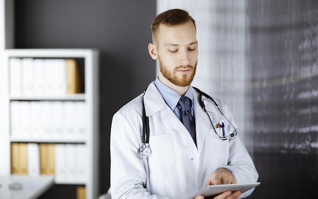 Sympathique médecin à barbe rousse utilisant un ordinateur tablette en clinique sur son lieu de travail. Service médical parfait dans le concept d'hôpital et de médecine.