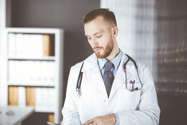 Sympathique médecin à barbe rousse utilisant un ordinateur tablette en clinique sur son lieu de travail. Service médical parfait dans le concept d'hôpital et de médecine.