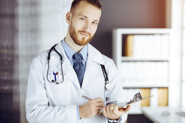 Sympathique médecin barbu debout et écrivant avec un presse-papiers dans une clinique ensoleillée sur son lieu de travail. Notion de médecine.