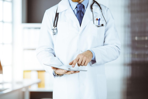 Sympathique médecin barbu utilisant une tablette dans une clinique ensoleillée sur son lieu de travail.