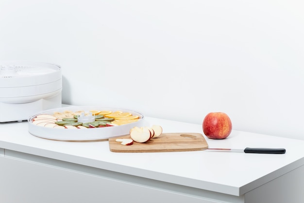 Sur une table blanche, il y a une pomme rouge, une planche à découper, un couteau de cuisine, un déshydrateur et un plateau avec des tranches de fruits.