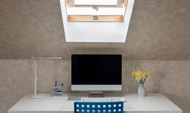 Une table blanche avec un moniteur, une lampe de table et un vase avec des jonquilles fraîches. Bureau à domicile