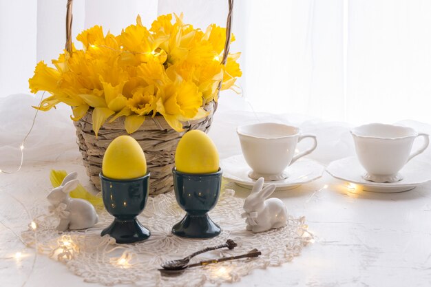 sur une table blanche des oeufs jaunes, des jonquilles dans un panier, deux tasses et des figurines en forme de lapins