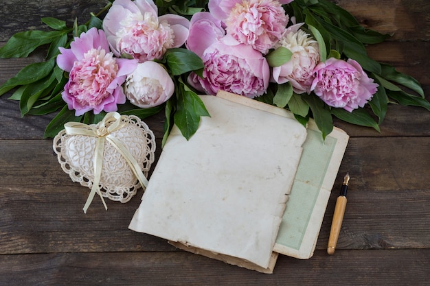 sur une table en bois un bouquet de pivoines roses, un stylo, des feuilles de vieux papier et un coeur