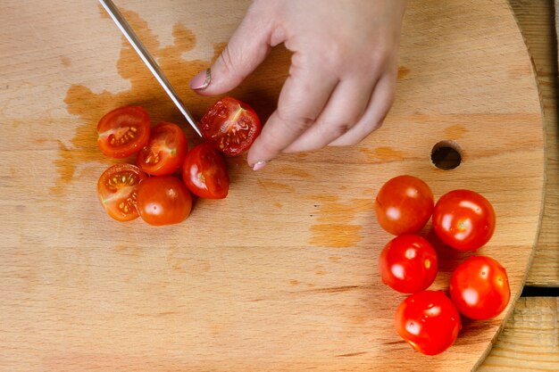 Sur une table en bois, une femme coupe des tomates cerises pour une salade de légumes avec un couteau.