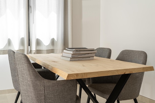 Table en bois marron clair avec des livres et quatre chaises à côté d'une fenêtre avec des rideaux