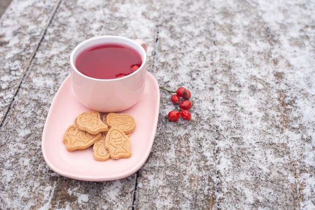 Sur une table en bois recouverte de givre se trouve une tasse de thé rose À proximité se trouvent des églantiers et des biscuits
