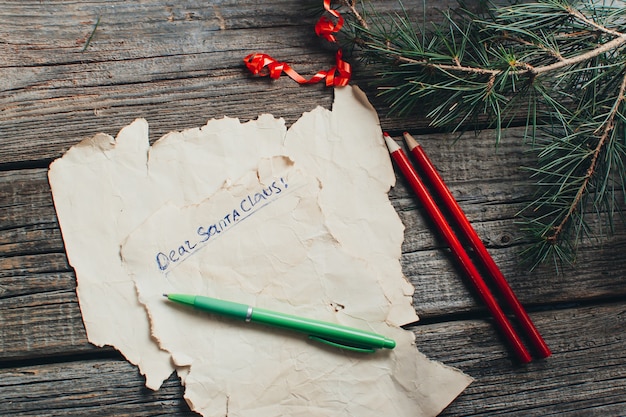 Sur la table en bois se trouve un vieux papier jaune qui dit, cher Père Noël rouge, crayons, stylos, cadeau de couleurs assorties et branches d'arbres pour la nouvelle année