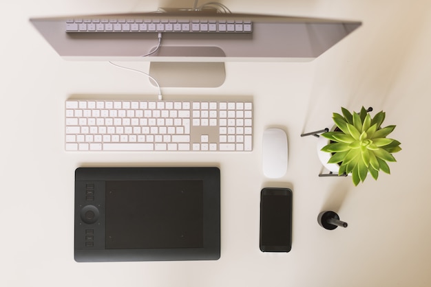 Table de bureau blanche avec clavier, souris, moniteur, tablette graphique, smartphone, plante succulente et autres fournitures de bureau.