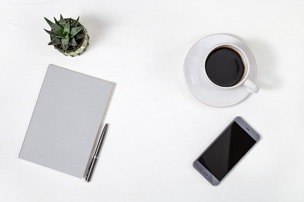 Table de bureau minimaliste blanche avec téléphone, cahier fermé gris, stylo et petite plante de cactus, tasse de café. Vue de dessus avec espace copie. Mise à plat.