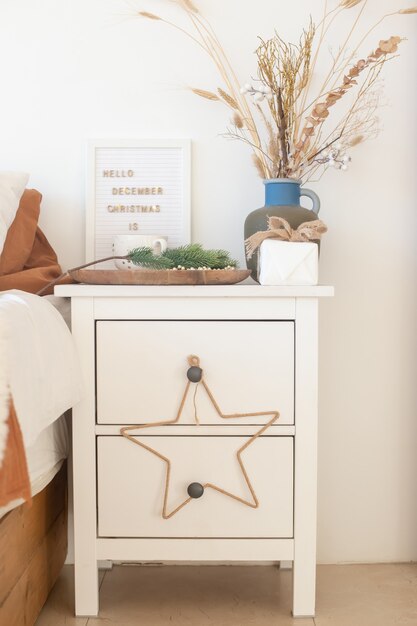 Table de chevet blanche avec décorations de Noël et fleurs séchées dans un vase bleu