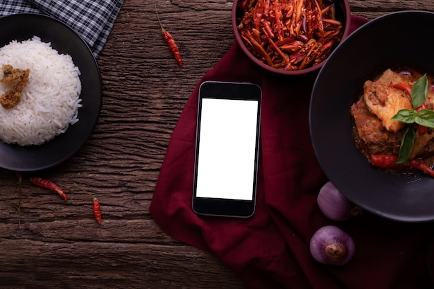 Photo table de cuisine avec écran blanc sur smartphone, tablette, téléphone portable et curry de porc rouge séché à la noix de coco.