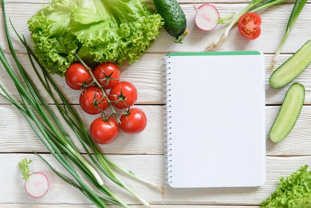 Table de cuisine rurale en bois blanc avec livre de cuisine vierge pour la liste de courses avec des ingrédients de salade fraîche (tomates, oignons, laitue, concombre). Livre de recettes vierge, espace de copie, vue de dessus