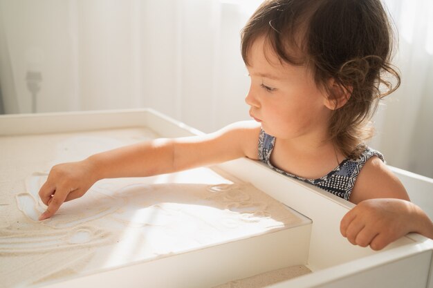 Table à dessin de sable pour enfants. Une petite fille dessine avec son doigt sur une table de sable clair