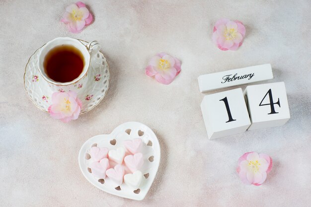 sur la table est une tasse de thé, des fleurs, des guimauves en forme de coeur et un calendrier avec la date du 14 février