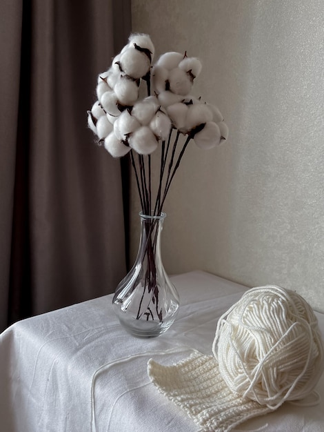 Photo sur la table il y a un vase avec des fleurs de coton et il y a un écheveau de laine blanche avec un carré tricoté commencé