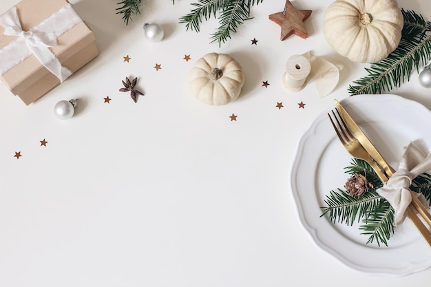 Table de Noël traditionnelle couverts dorés assiette en porcelaine branches de sapin boîte-cadeau pommes de pin et citrouilles blanches décoration de boules de Noël fond de vacances haut plat