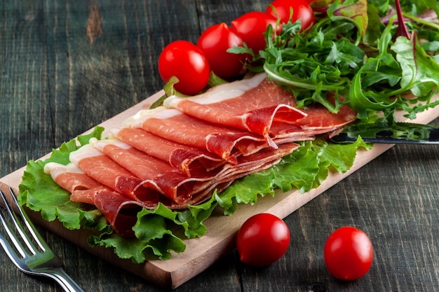 Sur la table se trouve une planche à découper en bois avec du jambon en tranches, des tomates et des herbes