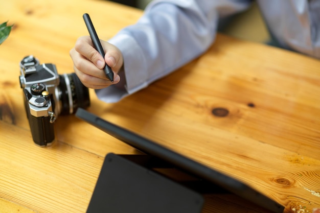 Table de travail en bois simple en gros plan avec appareil photo, tablette et main de femme tenant un stylet.