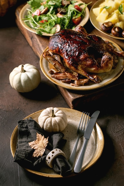 Table de vacances avec plats classiques canard glacé rôti aux pommes, pommes de terre bouillies, salade verte et sauce, assiette en céramique vide avec serviette et feuille d'automne sur table sombre avec décor d'automne.