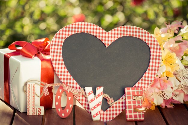 Tableau blanc en forme de coeur, boîte-cadeau et fleurs sur fond de printemps. Notion de fête des mères