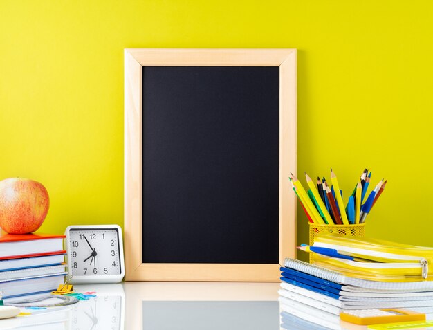 Tableau, pomme et fournitures scolaires sur une table blanche par le mur jaune. Vue de côté, vide