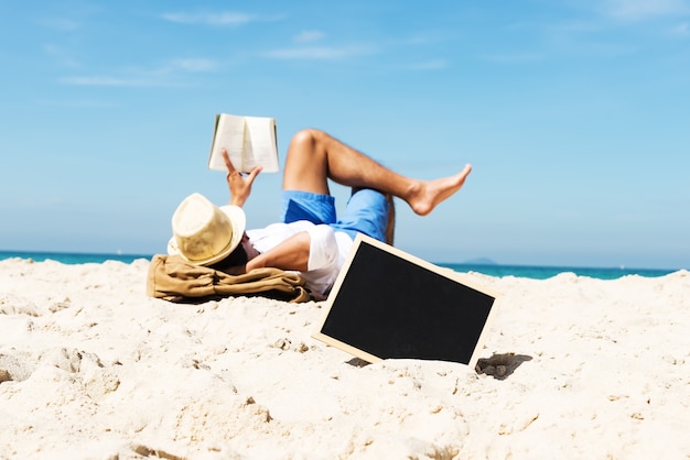 Tableau vide sur le sable de la plage avec jeune homme de touristes allongé sur la plage et lire un livre dans