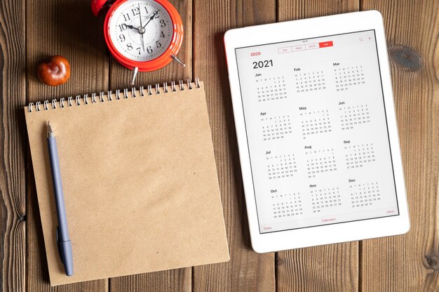 Une tablette avec un calendrier ouvert pour 2021 ans, un réveil rouge, des châtaignes et un cahier de papier craft sur un fond de table en bois