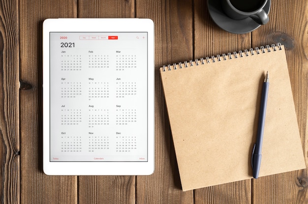 Une tablette avec un calendrier ouvert pour 2021 ans, une tasse de café et un cahier de papier craft sur un fond de table en bois