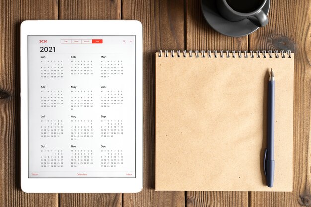 Une tablette avec un calendrier ouvert pour 2021 ans, une tasse de café et un cahier de papier craft sur un fond de table en bois