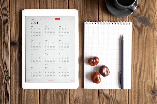 Une tablette avec un calendrier ouvert pour 2021 ans, une tasse de café, des châtaignes et un cahier de printemps avec un stylo sur un fond de table en bois