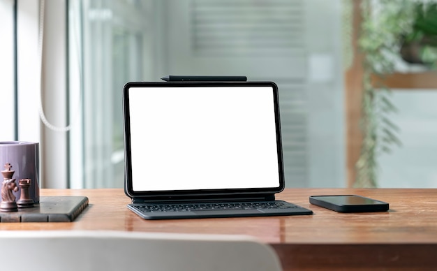 Tablette à écran blanc avec clavier magique et smartphone sur table en bois dans le salon.