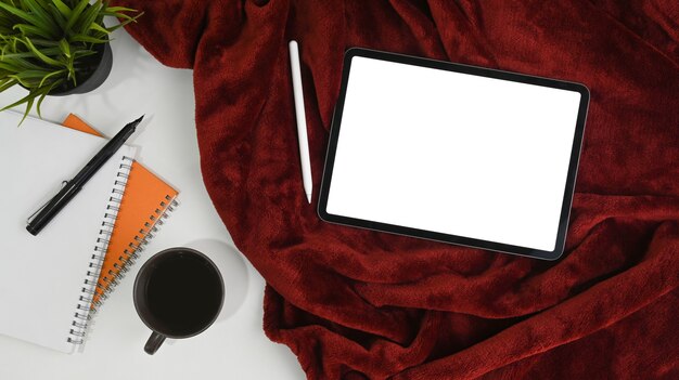 Tablette numérique avec écran blanc, stylet, tasse à café et bloc-notes sur couverture rouge.