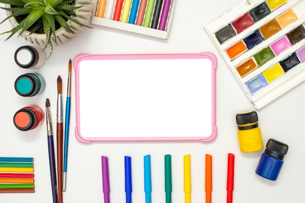 Tablette numérique rose pour enfants avec un écran vide sur un tableau blanc avec des fournitures de dessin multicolores