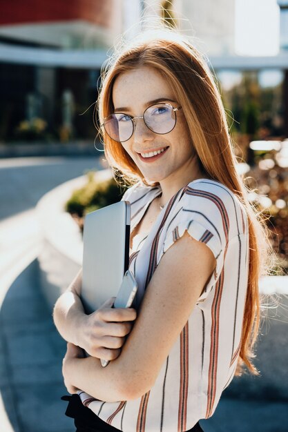 Taches de rousseur femme caucasienne aux cheveux rouges embrassant un ordinateur et regardant la caméra à travers des lunettes