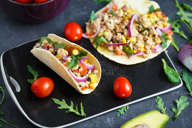 Tacos mexicains au porc, légumes et épices sur une assiette en pierre noire sur un fond sombre avec des ingrédients pour les tacos