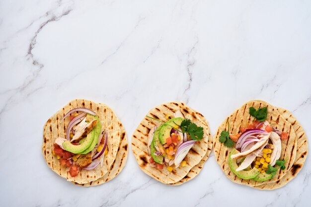 Tacos mexicains avec poulet grillé, avocat, grains de maïs, tomate, oignon, coriandre et salsa à table en pierre blanche. Cuisine de rue traditionnelle mexicaine et latino-américaine. Vue de dessus.