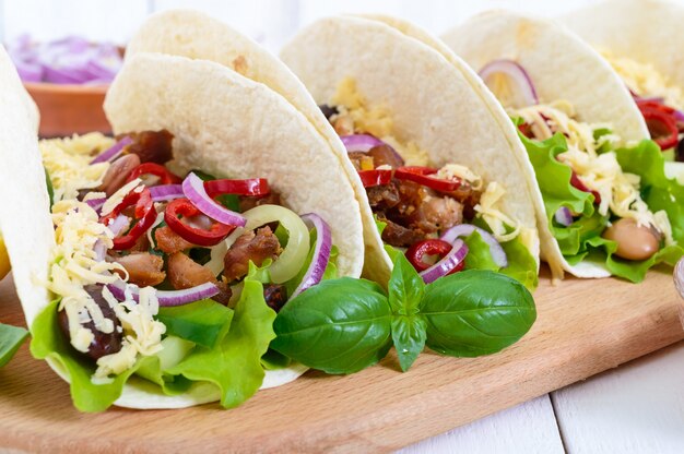 Les Tacos Sont Un Plat Mexicain Traditionnel. Tortilla Farcie Au Poulet, Cloche Et Piments Forts, Haricots, Laitue, Fromage, Oignon Bleu Avec Sauce Salsa Sur Fond De Bois Blanc.