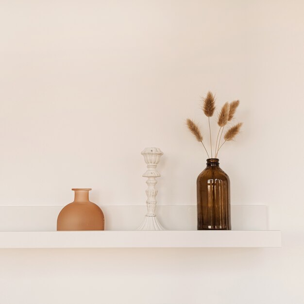 Étagère blanche sur mur blanc avec bouteille, vase, plantes à pompons moelleux, chandelier