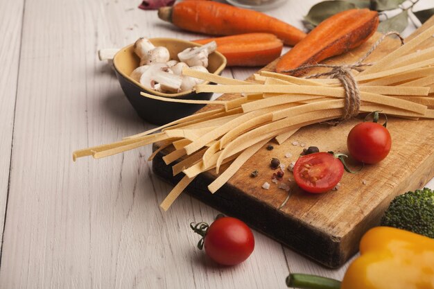 Tagliatelles crues faites à la main et divers légumes sur une table en bois blanc. Fond de pâtes de cuisson