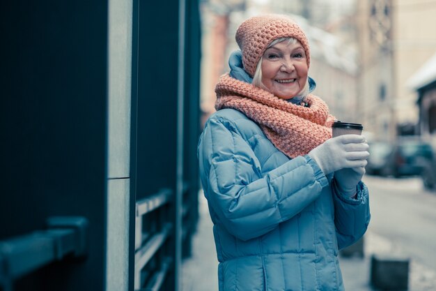 Taille d'une femme âgée positive debout dans la rue avec une tasse de café et souriant. Vêtements d'hiver chauds sur la femme