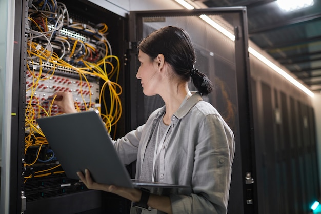 Taille d'une ingénieur réseau féminin connectant les câbles dans l'armoire du serveur tout en travaillant avec un superordinateur dans un centre de données, espace de copie