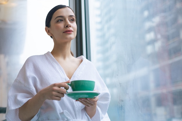 Taille d'une jeune femme calme vêtue d'un peignoir blanc tout en se tenant seule près de la fenêtre avec une tasse dans les mains