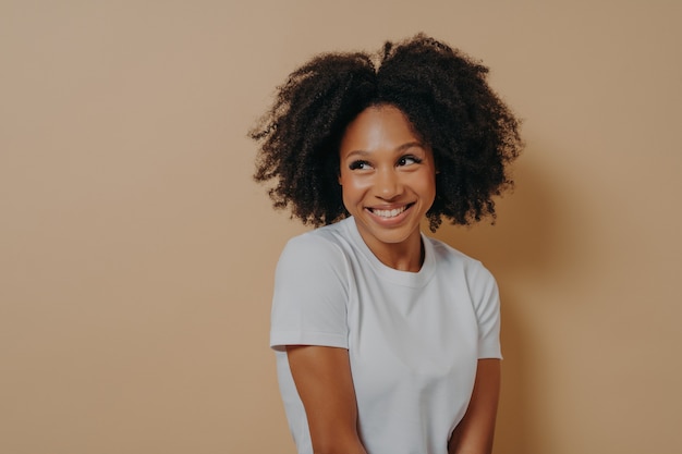 Taille d'une jolie femme africaine bouclée avec un sourire à pleines dents vêtue d'un t-shirt blanc regardant de côté avec timidité et exprimant des émotions positives, isolée sur fond beige. Concept d'expressions de visage