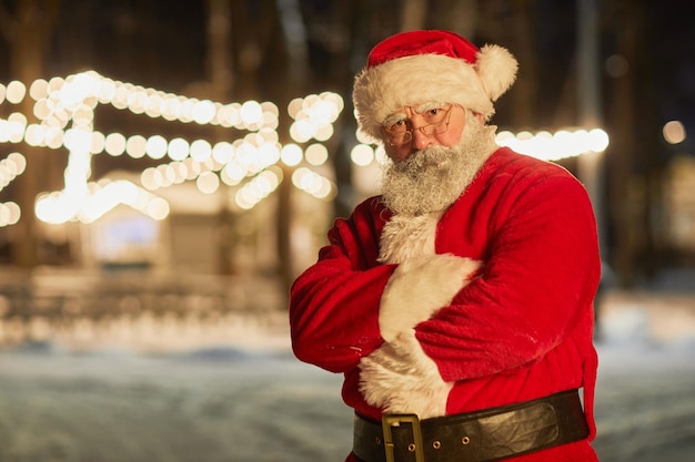 Taille portrait du père noël regardant la caméra tout en se tenant à l'extérieur la nuit avec les bras croisés