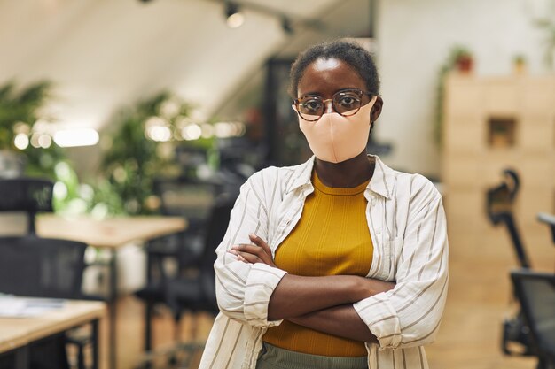 Taille portrait de femme afro-américaine confiante portant un masque au bureau et debout avec les bras croisés au bureau, copiez l'espace