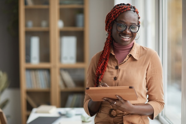 Taille portrait de femme afro-américaine contemporaine tenant une tablette numérique et souriant joyeusement en se tenant debout par la fenêtre au bureau, copiez l'espace