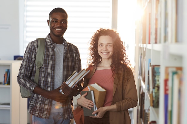 Taille portrait d'homme afro-américain debout dans la bibliothèque du collège avec jeune femme, à la fois tenant des livres et souriant à la caméra